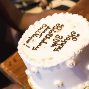 Chocolate cake with strawberry jam layers, covered in white buttercream and decorated with gold sparkle and chocolate lettering.