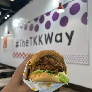 Holding chicken sandwich deluxe with poster behind saying "#TheTKKWay."