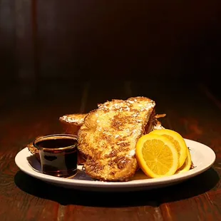 Thick sliced challah break French toast, stacked with orange slices and topped with powdered sugar. Served with a side of maple syrup.
