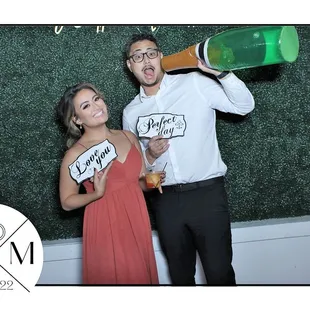 Posing in front of our Magic Mirror Photo Booth. Green Box hedge backdrop, photo booth props, neon sign, custom template, wedding event.