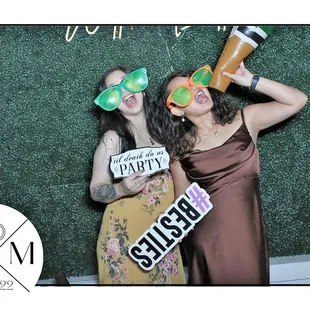 Posing in front of our Magic Mirror Photo Booth. Green Box hedge backdrop, photo booth props, neon sign, custom template, wedding event.