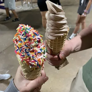 Small waffle cone - one with strawberry ice cream and sprinkles. The other with chocolate ice cream
