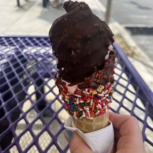 Strawberry soft serve ice cream dipped in chocolate with a waffle rainbow sprinkled cone.