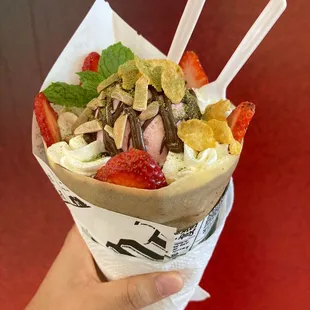 a hand holding a paper cone with strawberries, strawberries, and other toppings