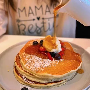 a woman pouring syrup over a stack of pancakes