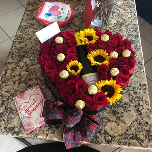 A roses and sunflower filled heart shape pot with wine bottle and chocolates.