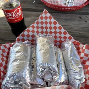 three burritos wrapped in aluminum foil on a picnic table