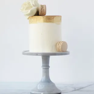 A simple white cake, decorated with a gold band, fresh white Roses, and vanilla macarons.