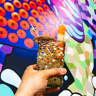 a hand holding a drink in front of a colorful mural