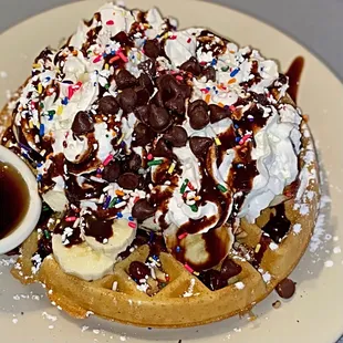 Chocolate chip pecan waffle topped with powdered sugar, bananas, whipped cream, chocolate sauce, and rainbow sprinkles.
