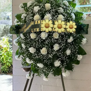 Stand spray with white carnation and a few roses.  Wording in gold on a green banner/ribbon.