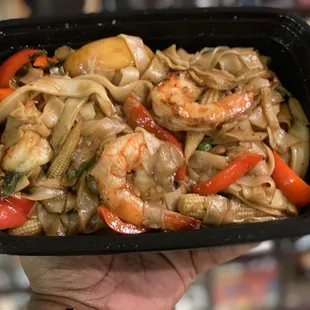 a hand holding a black plastic container filled with noodles, shrimp, and vegetables