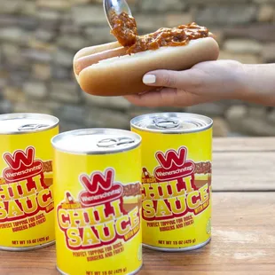 a person holding a hot dog and three cans of chili sauce