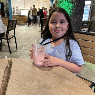 a little girl sitting at a table with a drink