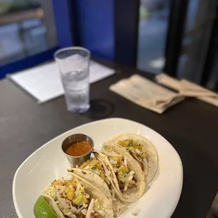 three tacos on a white plate with a glass of water