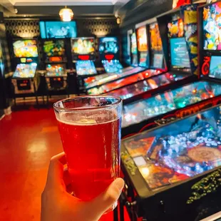 a person holding a glass of beer in front of a row of pinball machines