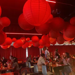a large group of people sitting at tables with red paper lanterns hanging from the ceiling