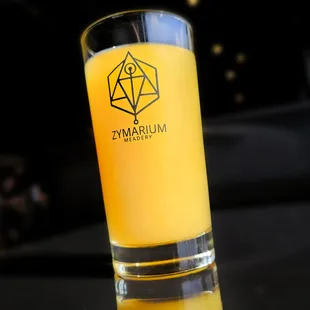 Orange mead in a tall glass with Zymarium Meadery logo on the glass. The taproom is faintly visible in the background